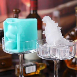 May include: A teal silicone ice mould and a clear glass filled with ice cubes, with a Godzilla-shaped ice sculpture. The ice sculpture is in a clear glass. The background includes bottles of liquor.