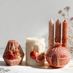 May include: A collection of decorative wax candles including a dusty rose pentagonal candle with a cathedral relief, a cream cylindrical candle with a pomegranate motif, three tall tapered candles with pomegranate designs, and a round pomegranate candle.