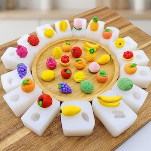 Könnte beinhalten: Eine Sammlung farbenfroher, miniaturförmiger Obstformen, darunter Bananen, Trauben und Erdbeeren, angeordnet auf einem Holztablett und einem Holzschneidebrett. Die Formen sind weiß und die Früchte sind hell gefärbt.