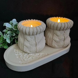 Puede incluir: Dos portavelas de cerámica blanca con forma de suéteres de punto con velas encendidas en su interior, colocados sobre una bandeja de cerámica blanca con un diseño decorativo.
