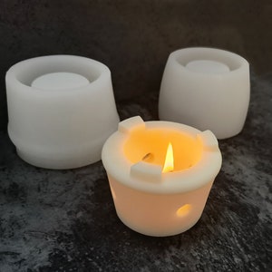 May include: Three white ceramic candle holders. One holder has a lit candle inside. The candle holder with the lit candle has a unique design with four holes around the top.