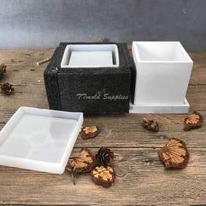 May include: A set of three white silicone molds for making square concrete planters. The molds are shown assembled and disassembled. The molds are on a wooden surface with pine cones and other natural elements.
