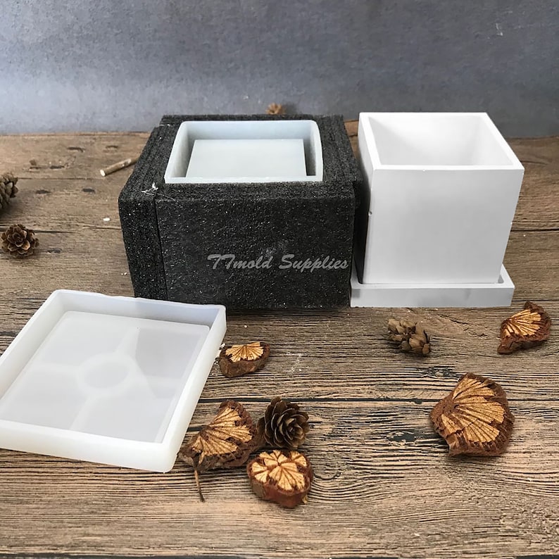 May include: A set of three white silicone molds for making square concrete planters. The molds are shown assembled and disassembled. The molds are on a wooden surface with pine cones and other natural elements.