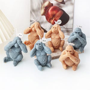 Può includere: Una collezione di sei candele a forma di scimmia nei toni del grigio e del beige. Ogni candela raffigura una scimmia seduta, con le mani che coprono gli occhi, le orecchie o la bocca. Piccole etichette bianche con la parola "Smile" sono attaccate.