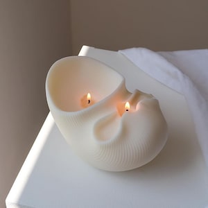 Op de afbeelding: Een witte kaars in de vorm van een gezicht met twee brandende lonten. De kaars heeft een getextureerd oppervlak.