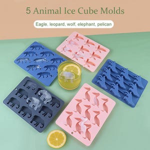 Puede incluir: Moldes para cubitos de hielo con cinco animales: águila, leopardo, lobo, elefante y pelícano. Los moldes son azules, rosas y grises. También se ve un vaso de limonada con una rodaja de limón y un cubito de hielo.