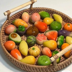 May include: A woven wicker tray holds an assortment of colorful, miniature fruit-shaped desserts. The treats include mangoes, watermelons, and other fruits, all with a glossy finish. The tray has wooden handles.