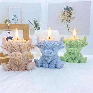 Pode incluir: Três velas esculpidas em forma de vacas Highland sentadas, cada uma com um pavio aceso no topo. As velas são de cor castanho-claro, azul suave e verde-pálido, apresentando texturas de pelo detalhadas, chifres curvos e olhos fechados.