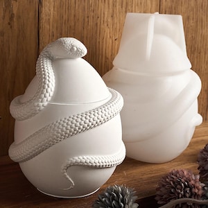 Może przedstawiać: Dwa białe ceramiczne pojemniki dekoracyjne. Jeden pojemnik ma kształt jajka z wężem owiniętym wokół niego. Drugi pojemnik ma kształt zaokrąglonej, abstrakcyjnej formy.