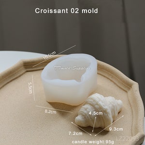 Puede incluir: Un molde de silicona blanco con forma de croissant, que mide 8,2 cm de ancho, 5,5 cm de alto y 10,5 cm de largo. Junto al molde hay una vela blanca con forma de croissant, que mide 7,2 cm de largo, 4,5 cm de ancho y 9,3 cm de alto. La vela pesa 95 gramos. El texto "Timold Supplies" y "LZ22093" es visible en la imagen.