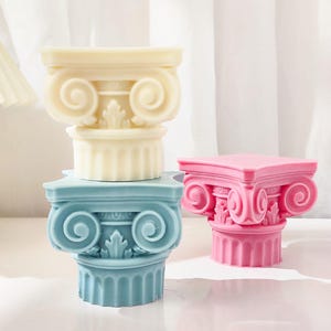 K&ouml;nnte beinhalten: Drei dekorative Kerzenhalter in Form von ionischen S&auml;ulen in Pastellfarben: Creme, Blau und Rosa. Jede S&auml;ule hat detaillierte Schn&ouml;rkel und eine quadratische Oberseite. Die S&auml;ulen sind &uuml;bereinander gestapelt.