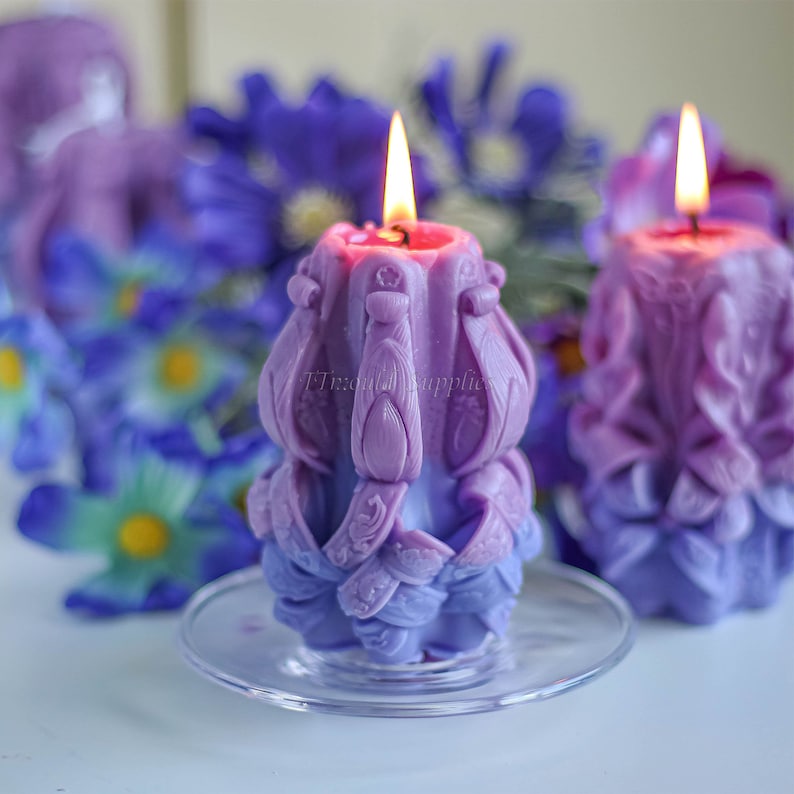May include: Two purple and lavender candles with intricate designs are lit and placed on a clear glass plate. The candles are surrounded by purple flowers.