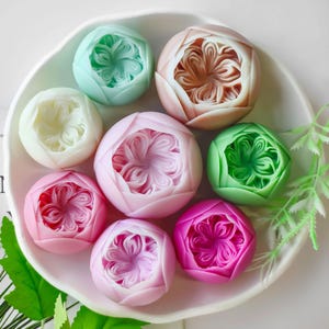 May include: A collection of seven handmade soap bars in various pastel colours, each with a detailed floral design. The soaps are arranged on a white plate.