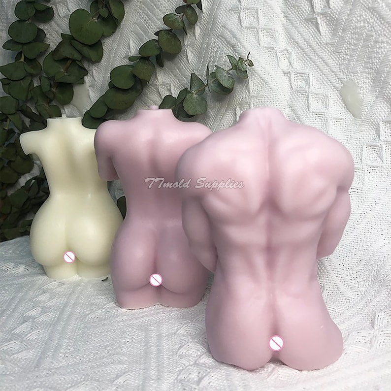 Op de afbeelding: Twee naaktkleurige kaarsen in de vorm van menselijke torso's. De kaarsen zijn gemaakt van een glad, zacht materiaal en hebben een realistisch uiterlijk. De kaarsen staan ​​naast elkaar op een wit oppervlak.
