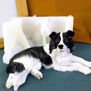 May include: A black and white border collie dog figurine lying down on a white surface. The dog has a black nose and black eyes. The figurine is made of a smooth, white material.