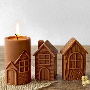 Può includere: Candela marrone con fiamma accesa e due candele a forma di casa. La candela presenta disegni a forma di casa. La candela e le case sono marroni, creando un'estetica rustica. La candela è cilindrica e le case hanno dettagliate texture di mattoni e legno.
