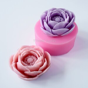 May include: Two silicone molds for making flower-shaped soap or candles. One mold is filled with a purple flower, and the other mold is next to a pink flower.