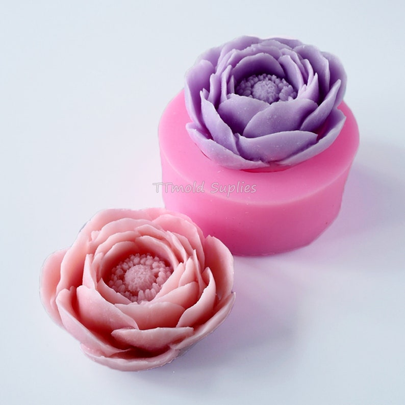 May include: Two silicone molds for making flower-shaped soap or candles. One mold is filled with a purple flower, and the other mold is next to a pink flower.