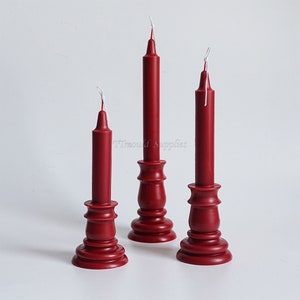 May include: Three red candles on wooden candle holders. The candles are tall and thin, with white wicks. The candle holders are a deep red color and have a classic design.