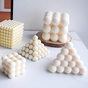 Puede incluir: Cinco esculturas de velas blancas en diferentes formas geométricas. Las velas están hechas de un material texturizado que se asemeja a pequeñas esferas. Las velas están dispuestas sobre una superficie blanca.