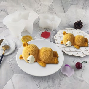 May include: Two white silicone molds shaped like sleeping bears. Two yellow sleeping bear cakes are on white plates.