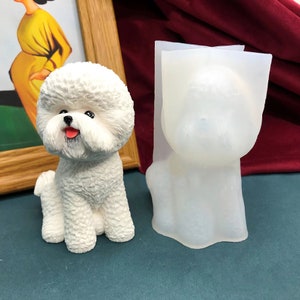 Puede incluir: Una figura de perro Bichon Frise blanco con un pelaje texturizado y esponjoso, ojos negros y una lengua rosa. Junto a ella, un molde de silicona transparente con la forma del perro. El fondo incluye un cuadro enmarcado y una tela burdeos.