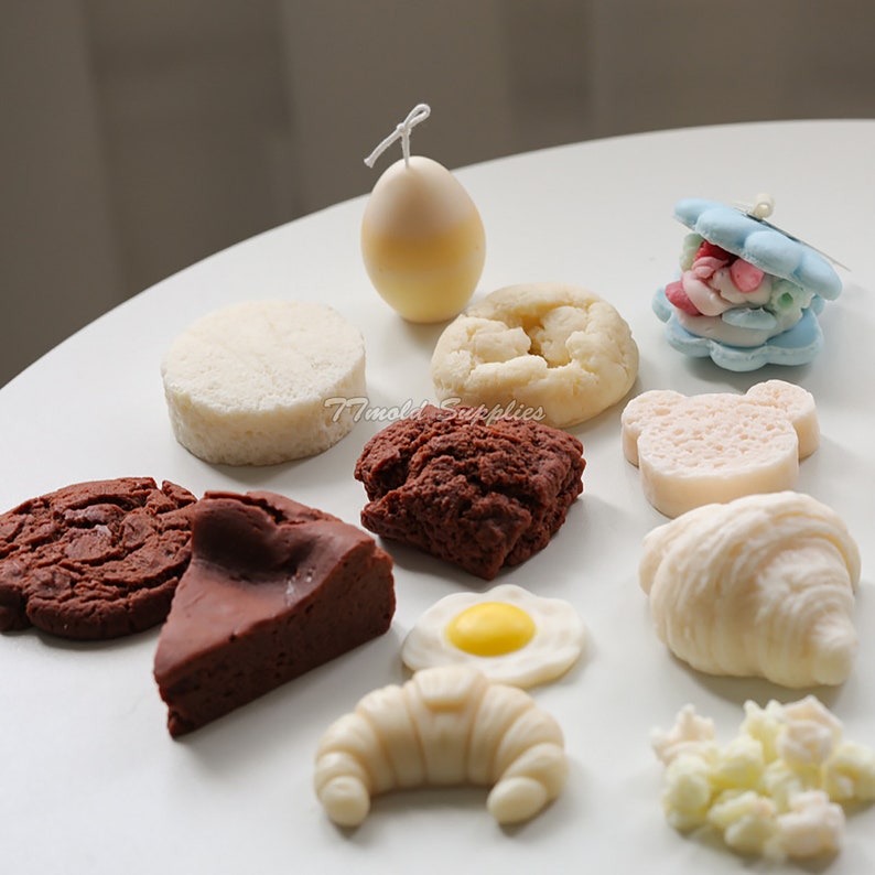 Puede incluir: Surtido de velas artesanales con forma de diferentes alimentos, incluyendo un croissant, una galleta, un huevo y una concha de almeja llena de caramelos coloridos. Las velas son blancas, marrones, amarillas y azules.