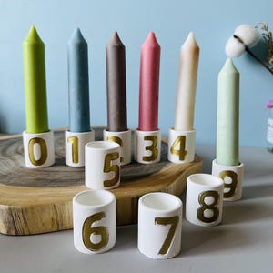 Puede incluir: Colección de velas coloridas en soportes de cerámica blanca con números dorados. Las velas son en tonos verdes, azules, marrones, rosas y beige. Los soportes están numerados del 0 al 9, ideales para un cumpleaños o aniversario.