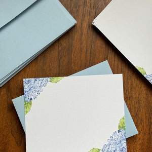 May include: A stack of light blue envelopes with a white card featuring a watercolor illustration of blue hydrangeas in the corners.