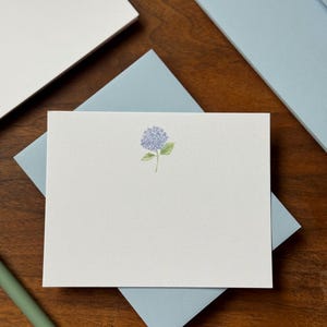 May include: Stationery set featuring a white card with a watercolor hydrangea illustration, resting on a light blue envelope. Additional white cards and light blue envelopes are visible, along with a green pen. The set is arranged on a wooden surface.