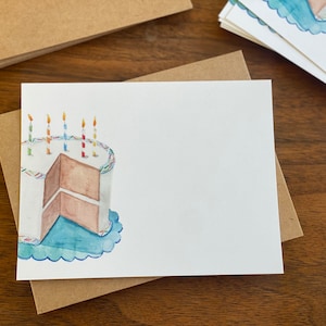 May include: A white card featuring a watercolour illustration of a birthday cake with candles. The cake is brown with white icing and blue sprinkles. The cake is on a blue plate with a scalloped edge.