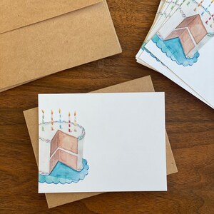 May include: A set of blank white cards featuring a watercolour illustration of a birthday cake with candles. The cake is brown with white icing and a blue plate. The cards are packaged with brown envelopes.