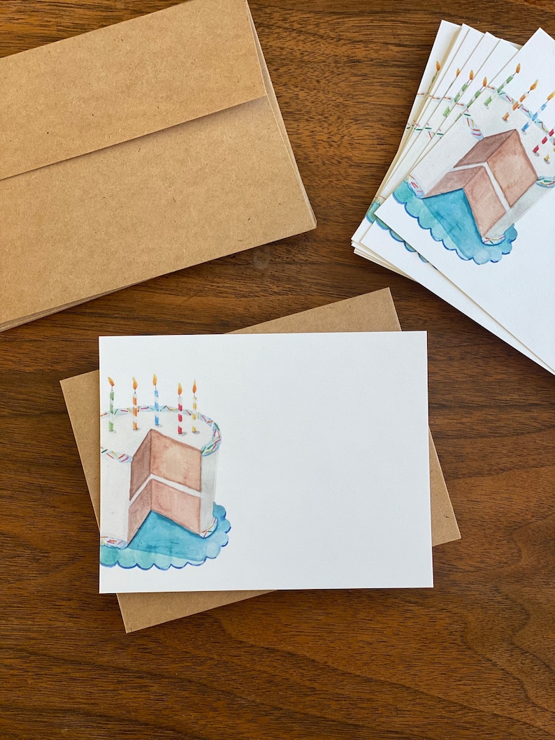 May include: A set of blank white cards featuring a watercolour illustration of a birthday cake with candles. The cake is brown with white icing and a blue plate. The cards are packaged with brown envelopes.