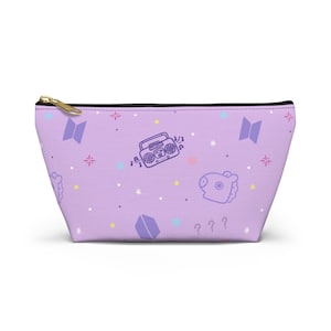 May include: A lavender cosmetic bag with a gold zipper. The bag features a repeating pattern of a retro radio, stars, hearts, and a cartoon character. The bag is designed with a black trim.