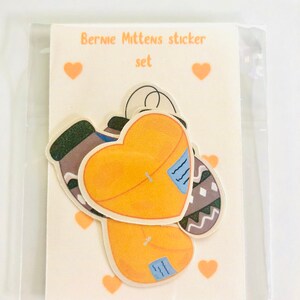 May include: A set of Bernie Mittens stickers featuring a heart-shaped design with a brown and white patterned mitten on each side. The stickers are on a white card with the text "Bernie Mittens sticker set" and hearts.
