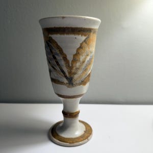 May include: A handmade ceramic goblet with a white base and a brown rim. The goblet features a leaf-like design in shades of brown, blue, and tan. The stem and base are also white with brown accents. The goblet is approximately 6 inches tall.