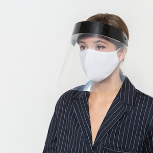 May include: A person wearing a white face mask and a clear plastic face shield with a black headband. The person is wearing a black and white striped blazer.
