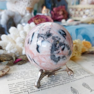 May include: A pink and black sphere with a swirling pattern, sitting on a silver stand. The sphere is made of polished stone and has a smooth, glossy finish.