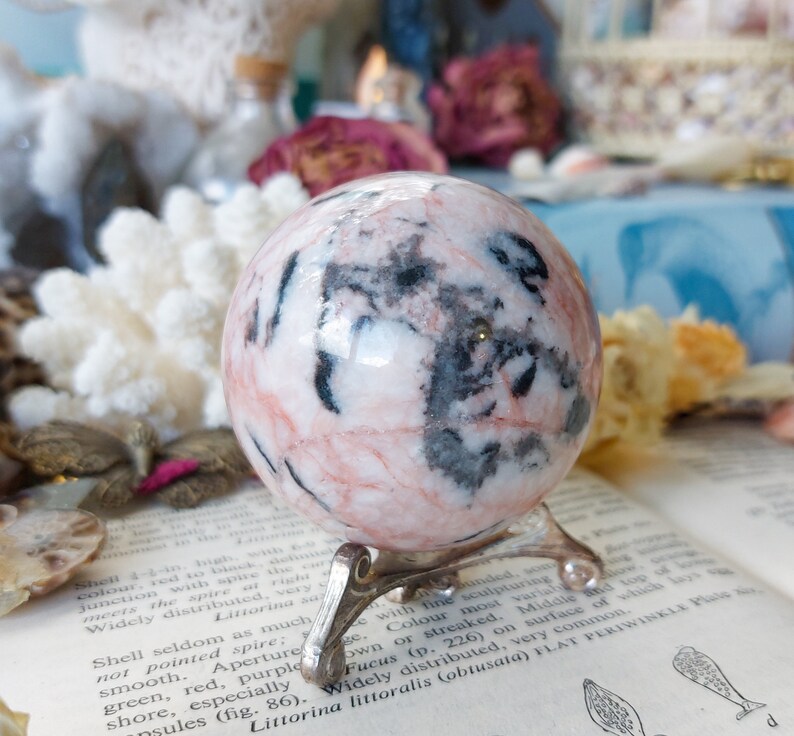 May include: A pink and black sphere with a swirling pattern, sitting on a silver stand. The sphere is made of polished stone and has a smooth, glossy finish.