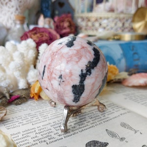 May include: A pink and black sphere with a swirling pattern, sitting on a metal stand. The sphere is made of polished stone and has a smooth, glossy finish.