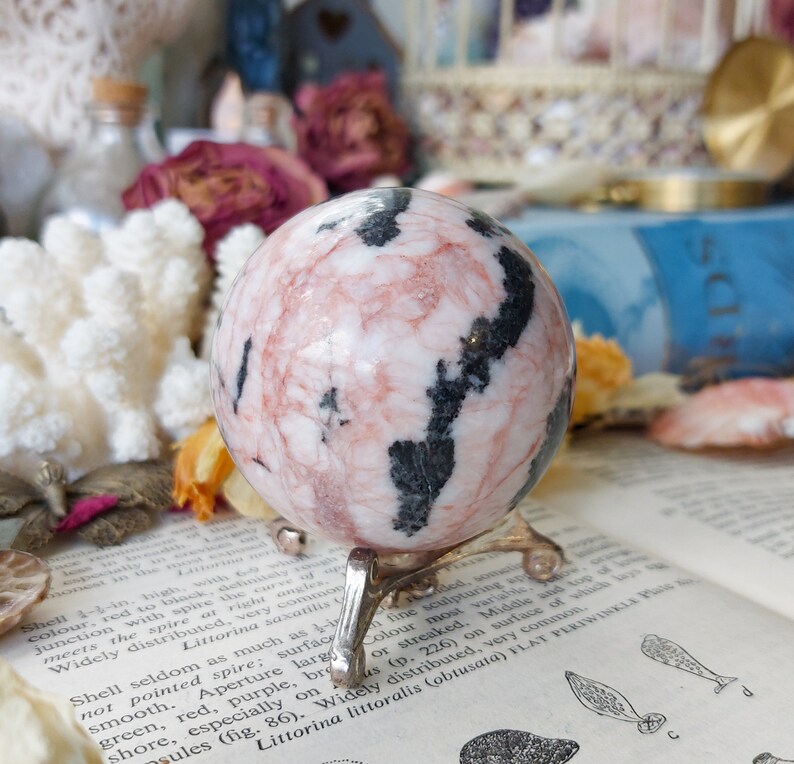 May include: A pink and black sphere with a swirling pattern, sitting on a metal stand. The sphere is made of polished stone and has a smooth, glossy finish.