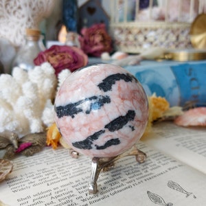May include: A pink and black zebra jasper sphere on a metal stand. The sphere is polished and has a smooth surface. The sphere is approximately 2 inches in diameter.