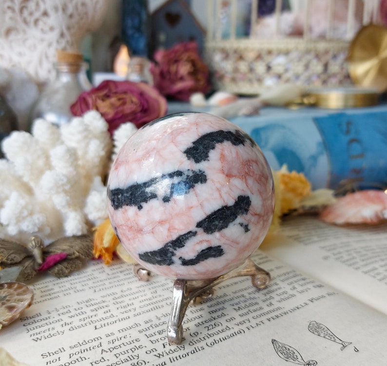 May include: A pink and black zebra jasper sphere on a metal stand. The sphere is polished and has a smooth surface. The sphere is approximately 2 inches in diameter.