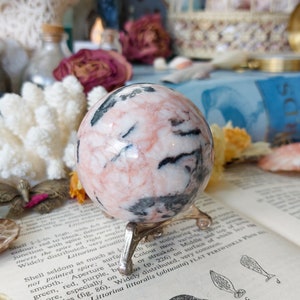 May include: A pink and black sphere of polished stone on a gold metal stand. The sphere has a swirling pattern of pink, white, and black.