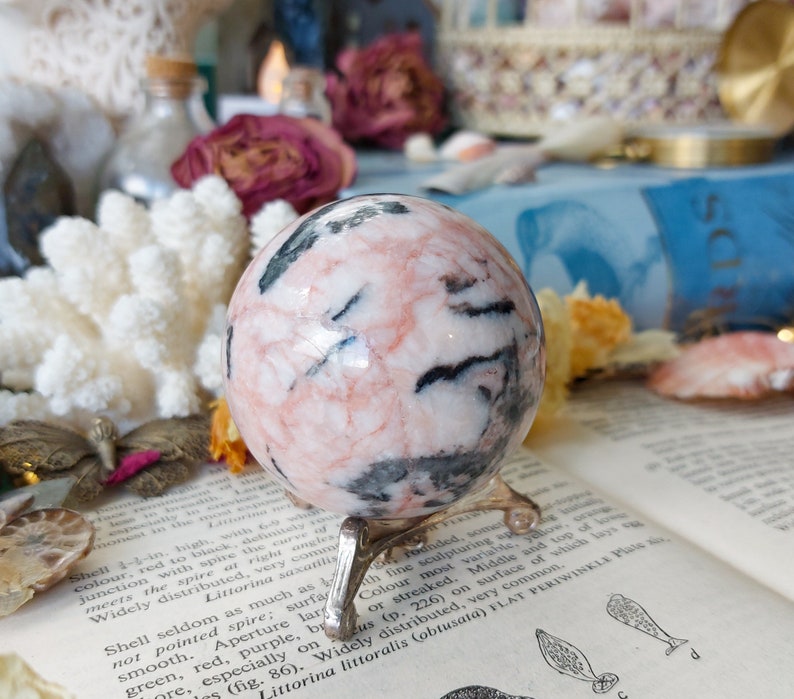 May include: A pink and black sphere of polished stone on a gold metal stand. The sphere has a swirling pattern of pink, white, and black.