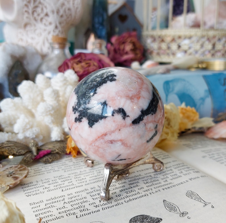 May include: A pink and black sphere of polished rhodochrosite, a type of mineral, on a silver stand. The sphere is about 2 inches in diameter.