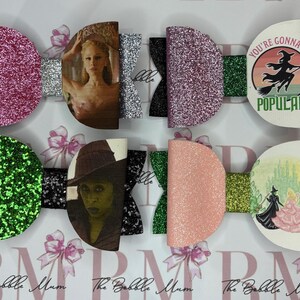 May include: A selection of decorative hair bows with glitter detailing and images from the musical "Wicked". The bows feature character images and the text "You're Gonna Be Popular". Each bow has a unique colour scheme.