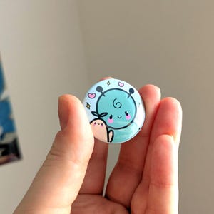 Könnte beinhalten: Ein runder, hellblauer Button mit einer Cartoon-Illustration eines lächelnden Gesichts, Herzen und einer kleinen Kreatur. Der Button wird in einer Hand gehalten, mit unscharfem Hintergrund. Ein niedliches Accessoire.