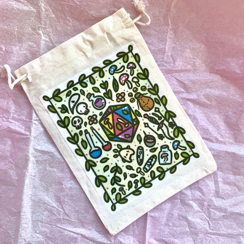 Puede incluir: Una bolsa de lona con cord&oacute;n que contiene una pegatina con ilustraciones de fantas&iacute;a. La pegatina presenta un dado D20, pociones, setas y una calavera, enmarcada por un borde de hojas verdes. La bolsa est&aacute; sobre una superficie rosa.
