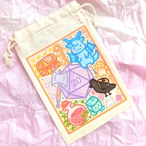 May include: A white drawstring bag with a colorful illustration of a 20-sided die, a dragon, a chest, an axe, a potion, a crow, and a mushroom. The bag is on a pink and white textured surface.